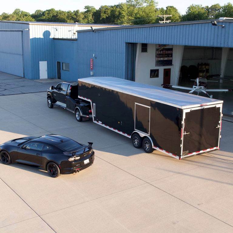A two-car enclosed auto transport trailer in black is being hauled by a black dually pickup truck.