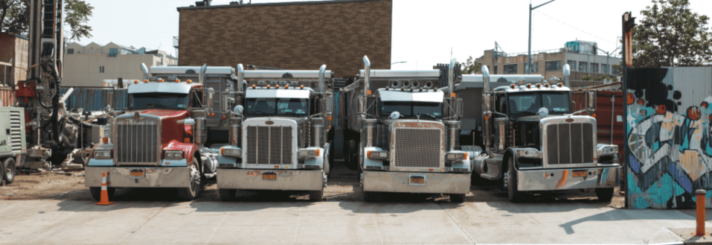 Four Semi-Trucks are parked in a dirt lot. We can ship a car with either an open car trailer or an enclosed car transport.